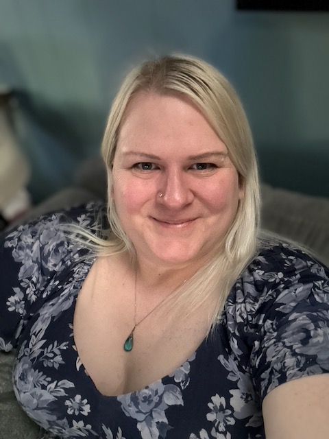 Blonde haired blue eyed author Chris Rue smiling at the camera in a floral dress and teal pendant